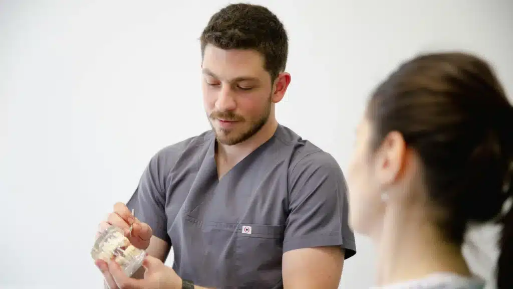 Male dentist in gray scrubs points to a model of teeth while explaining treatment to a patient in a dental office. This image conveys trust, care, and the importance of healthcare website development and custom website development services for premium practices.