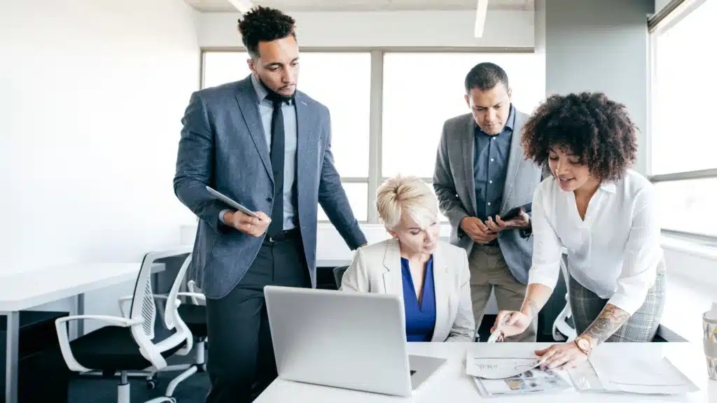 A professional marketing team in business attire gathers around a desk reviewing a printed strategy document. A woman leads the discussion, pointing to the paper as the team listens attentively. This scene illustrates collaboration typical of a top-tier Google Ads marketing agency or PPC consultant developing local SEO solutions for small businesses.