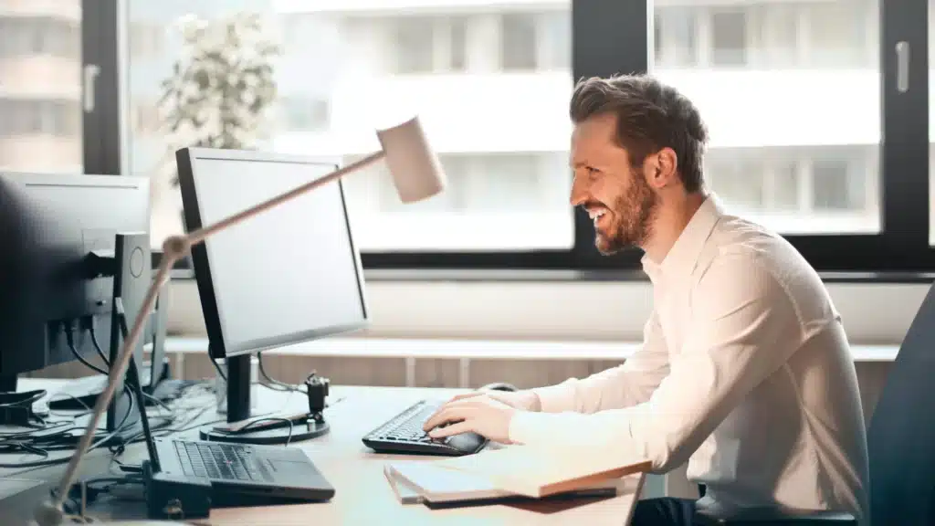 A smiling man working on a computer in a bright office setting. This image visually represents chiropractor marketing with a focus on digital marketing for chiropractors. The atmosphere is productive and positive, reflecting successful marketing ideas for chiropractors.